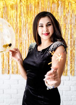 Birthday party. Beautiful young woman holding sparkler and balloon on golden background