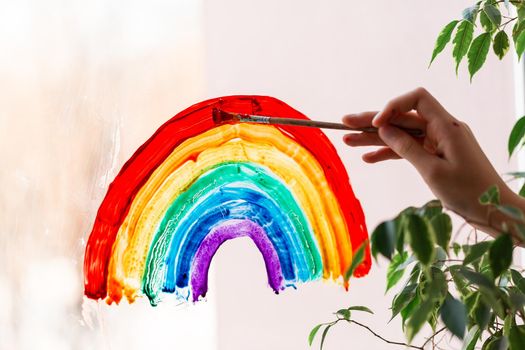 Little girl painting rainbow on window during Covid-19 quarantine at home. Stay at home social media campaign for coronavirus prevention, let's all be well, catch the rainbow