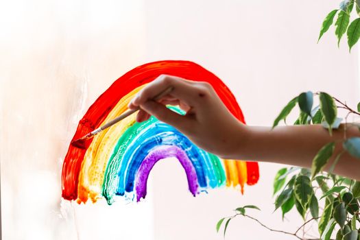 Little girl painting rainbow on window during Covid-19 quarantine at home. Stay at home social media campaign for coronavirus prevention, let's all be well, catch the rainbow
