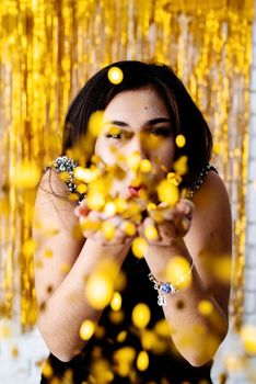 Birthday party. Portrait of young beautiful girl blowing golden confetti at holiday party