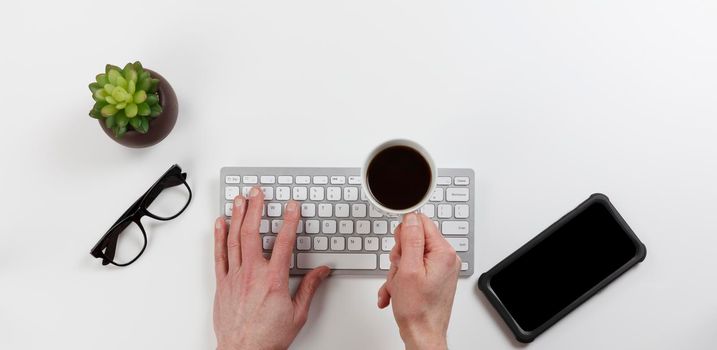 Office desk table with male hands holding cup of coffee over computer keyboard while working. Overhead with copy space available in flat lay format. 