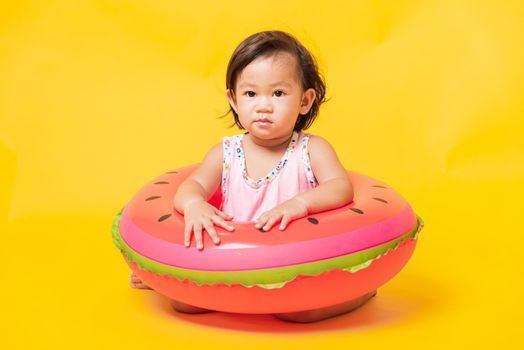 Summer vacation concept, Portrait Asian happy cute little baby girl wear swimsuit sitting in watermelon inflatable ring, Kid have fun sit in inflatable, studio shot isolated yellow background