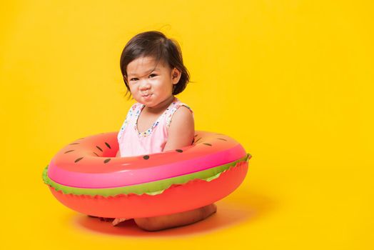 Summer vacation concept, Portrait Asian happy cute little baby girl wear swimsuit sitting in watermelon inflatable ring, Kid have fun sit in inflatable, studio shot isolated yellow background