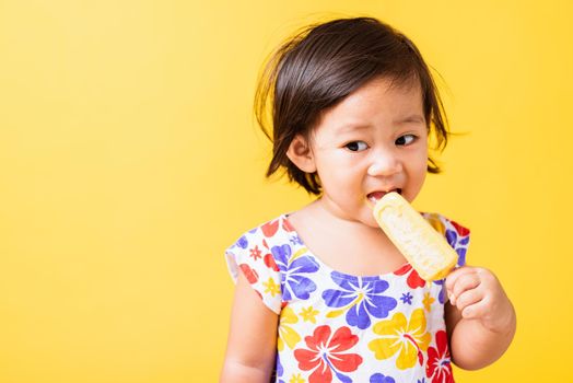 Happy portrait Asian baby or kid cute little girl attractive laugh smile wearing dick pattern shirt holds and eating sweet wooden ice cream, studio shot isolated on yellow background, summer concept