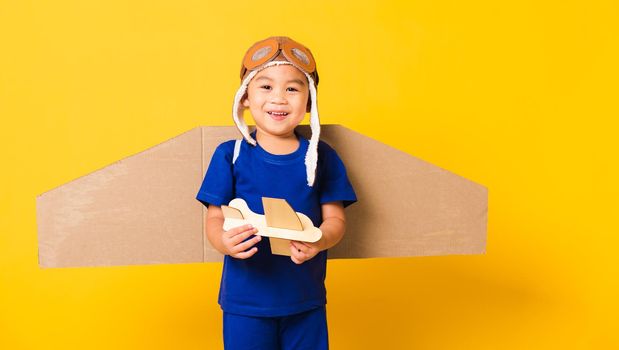 Happy Asian handsome funny child or kid little boy smile wear pilot hat play and goggles with toy cardboard airplane wings fly hold plane toy, studio shot isolated yellow background, Startup freedom