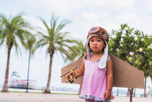 Happy Asian funny child or kid little girl smile wear pilot hat and goggles play toy cardboard airplane wing flying against summer sky cloud on trees garden background, Startup freedom concept