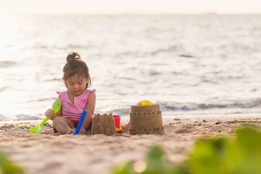 Happy fun Asian child cute little girl playing sand with toy sand tools at a tropical sea beach in holiday summer on sunset time, tourist trip concept