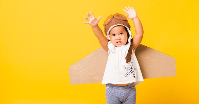 Happy Asian beautiful funny baby little girl smile wear pilot hat raise hand up play and goggles with toy cardboard airplane wings fly, studio shot isolated yellow background, Startup freedom concept