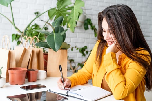 Home gardening. Small business. Young woman gardener making notes in the notepad surrounded with plants