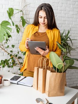 Home gardening. Small business. Young woman gardener making notes in the notepad, line of craft shopping bags for packing plant in the front