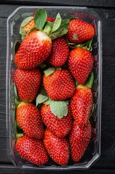 Red ripe strawberrie in clear plastic tray, on black wooden table background, top view flat lay