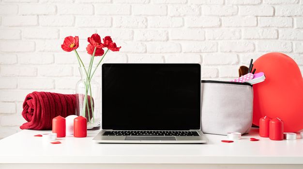 Valentines Day and Womens Day concept. laptop with cosmetic and spa accessories, gift box, flowers and candles on dressing table on white brick wall background