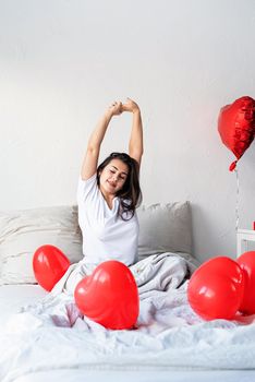 Valentine's Day. Sleeping. Young happy brunette woman sitting awake in the bed with red heart shaped balloons