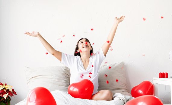 Valentine's Day. Sleeping. Young happy brunette woman sitting in the bed with red heart shaped balloons throwing confetti in the air