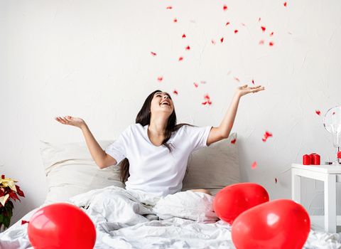Valentine's Day. Sleeping. Young happy brunette woman sitting in the bed with red heart shaped balloons throwing confetti in the air