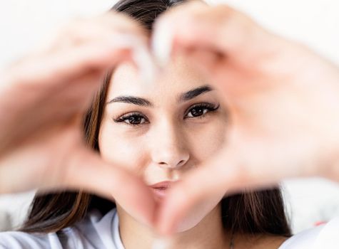 Valentine's Day. Young happy brunette woman in white t-shirt showing heart sign with her hands in front of face