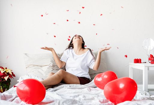 Valentine's Day. Sleeping. Young happy brunette woman sitting in the bed with red heart shaped balloons throwing confetti in the air