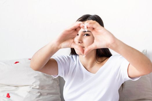 Valentine's Day. Young happy brunette woman in white t-shirt showing heart sign with her hands in front of face