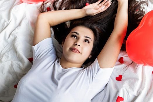 Valentines Day. Young happy brunette woman laying in the bed with red heart shaped balloons and decorations