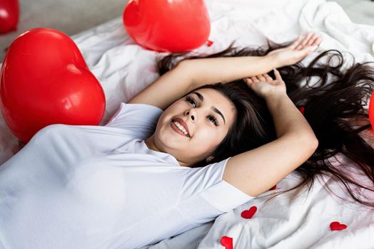 Valentines Day. Young happy brunette woman laying in the bed with red heart shaped balloons and decorations