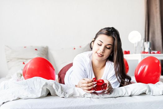 Valentine's Day. Sleeping. Young happy brunette woman laying in the bed with red heart shaped balloons wrapping the gift box