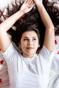 Valentines Day. Young happy brunette woman laying in the bed with red heart shaped confetti
