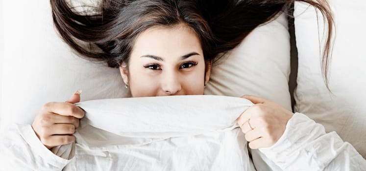young beautiful brunette woman sleeping in bed covering her face with blanket, top view flat lay