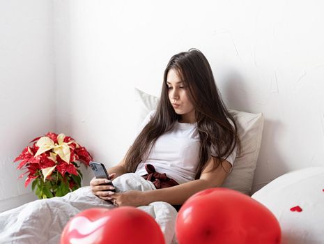 Valentines Day. Young brunette woman sitting awake in the bed with red heart shaped balloons and decorations texting