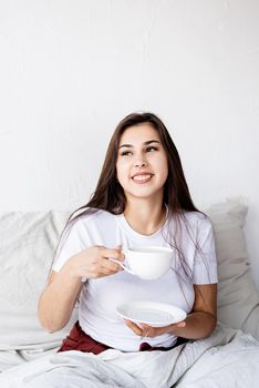 Valentines Day. Young brunette woman sitting awake in the bed with red heart shaped balloons and decorations drinking coffee eating croissants