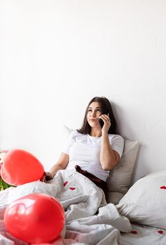 Valentines Day. Young brunette woman sitting awake in the bed with red heart shaped balloons and decorations texting