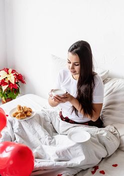 Valentines Day. Young brunette woman sitting awake in the bed with red heart shaped balloons and decorations drinking coffee eating croissants