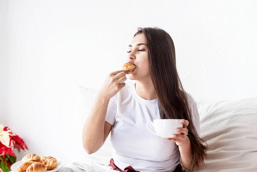 Breakfast and morning routine. Young brunette woman sitting in the bed with eating croissants