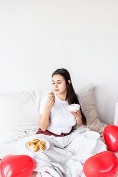 Valentines Day. Young brunette woman sitting awake in the bed with red heart shaped balloons and decorations drinking coffee eating croissants