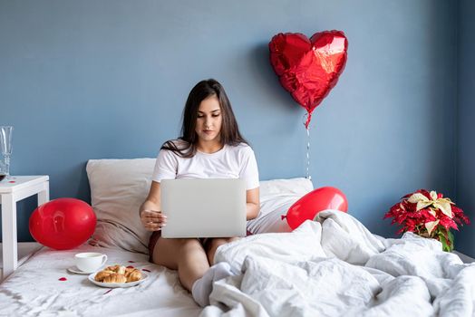 Valentine's Day. Young happy brunette woman sitting in the bed with red heart shaped balloons working on the laptop