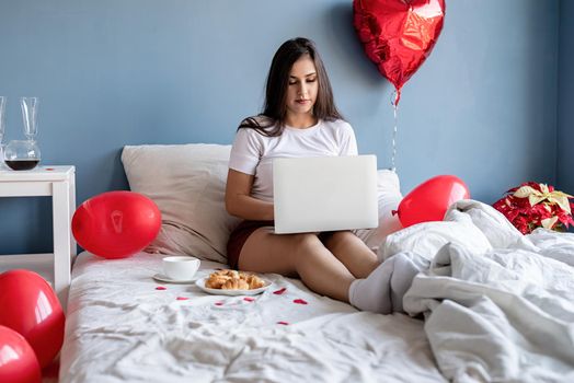 Valentine's Day. Young happy brunette woman sitting in the bed with red heart shaped balloons working on the laptop