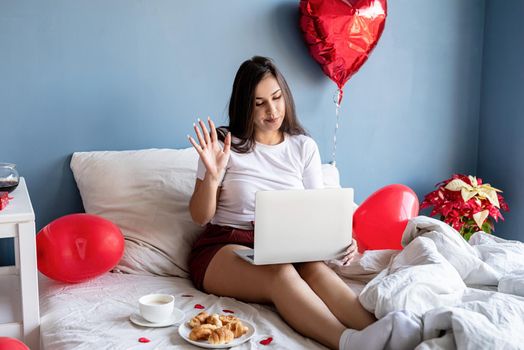 Valentine's Day. Young happy brunette woman sitting in the bed with red heart shaped balloons working on the laptop from home