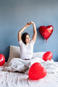 Valentine's Day. Sleeping. Young happy brunette woman sitting awake in the bed with red heart shaped balloons