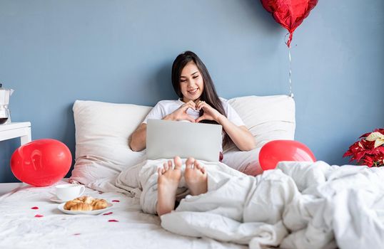 Valentines Day. Young happy brunette woman sitting in the bed with red heart shaped balloons chatting with her boyfriend on laptop showing heart gesture with hands