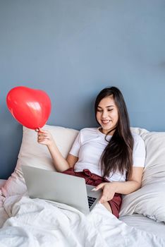 Valentine's Day. Young happy brunette woman sitting in the bed with red heart shaped balloons working on the laptop from home greeting friends