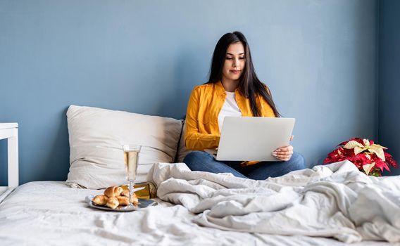 Valentines Day. Young brunette woman sitting in the bed working on computer drinking champagne
