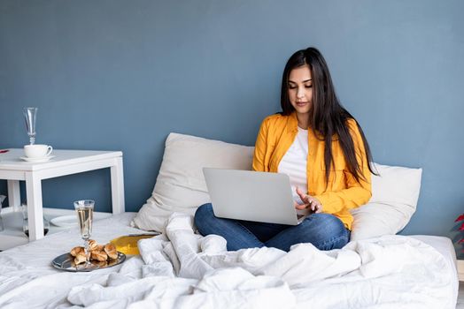 Valentines Day. Young brunette woman sitting in the bed working on computer drinking champagne