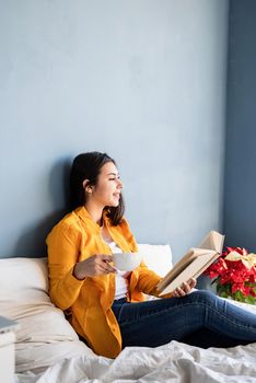 Breakfast and morning routine. Young brunette woman sitting in the bed with eating croissants and reading a book