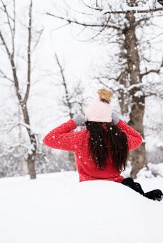 Winter season. Rear view of brunette woman sitting in snowy park in snowfall putting on her hat