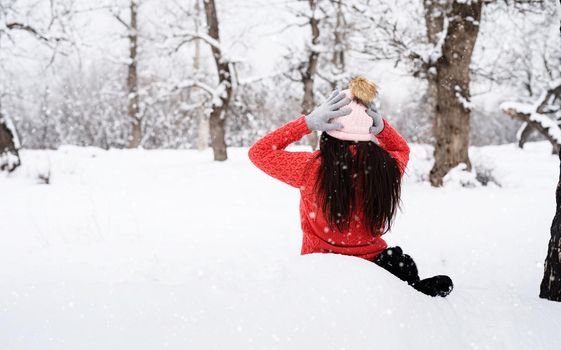 Winter season. Rear view of brunette woman sitting in snowy park in snowfall putting on her hat