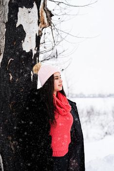 Winter season. Portrait of a beautiful smiling young woman standing by the tree in snowfall
