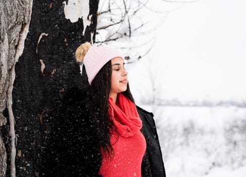 Winter season. Portrait of a beautiful smiling young woman standing by the tree with eyes closed in snowfall