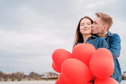 Valentines Day. Young loving couple hugging and kissing holding red heart shaped balloons outdoors