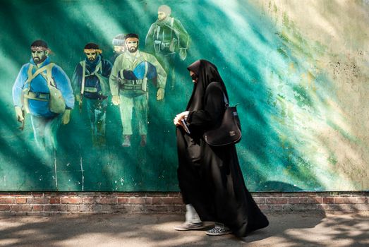 veiled muslim woman walking by revolutionary fighters mural in downtown tehran city street iran outside old US american embassy