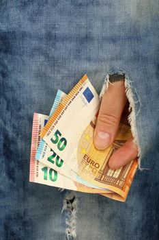 Hand holds several different value Euro paper currency banknotes in jeans rip hole, low angle view