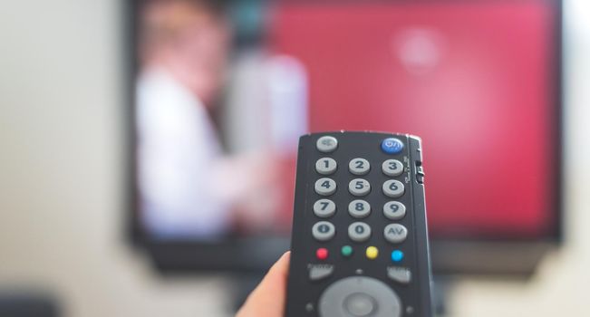 Holding a TV remote control in the hand, foreground, tv in the blurry background. Streaming.